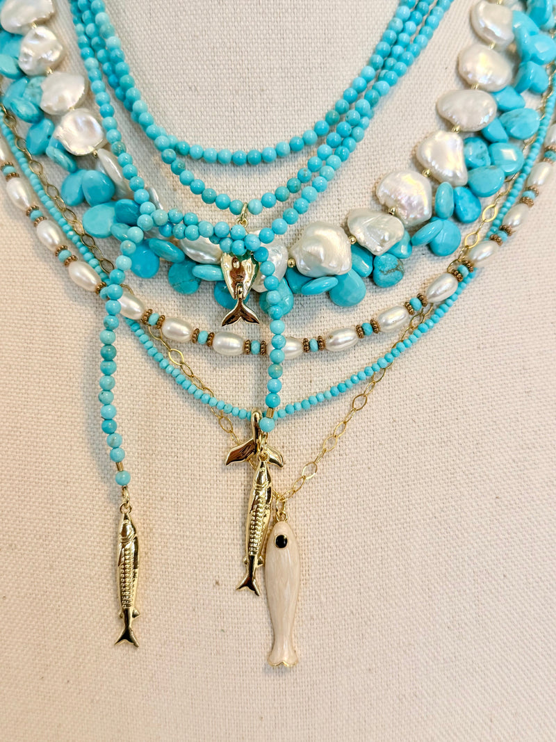 A multi-strand necklace displayed on a beige fabric mannequin. The necklace features multiple strands of turquoise beads, white baroque pearls, gold-tone chain links, and three dangling fish charms in gold-tone metal and white bone.
