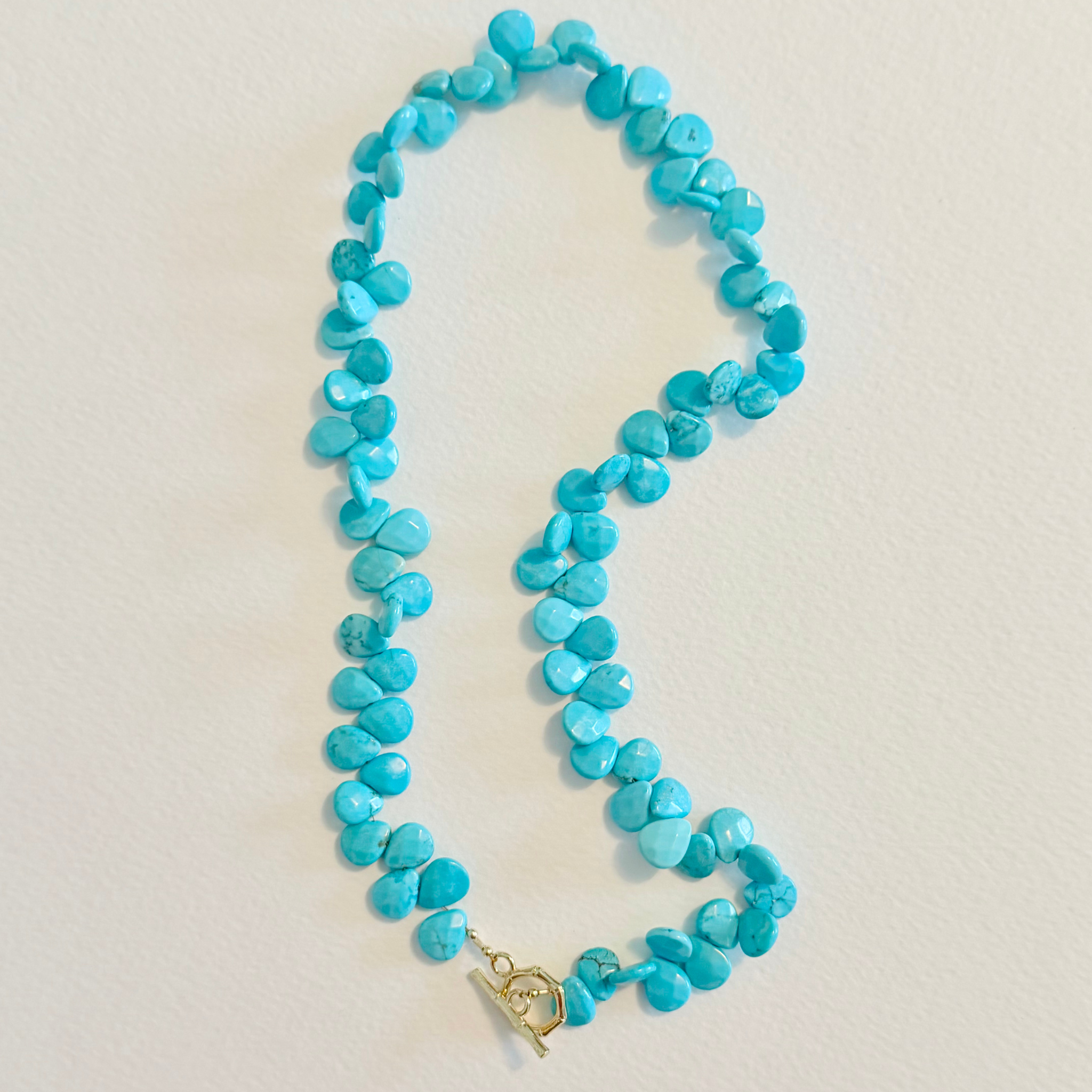 A turquoise stone bead necklace with a gold toggle clasp, laid out on a light beige textured surface.