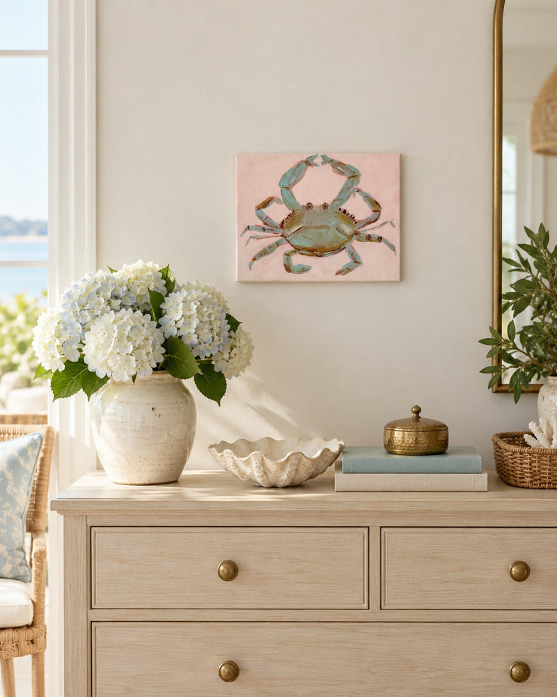 A rectangle canvas art print featuring a blue-green crab on a soft pink background, styled on a light wood dresser in a bright, coastal-themed room.
