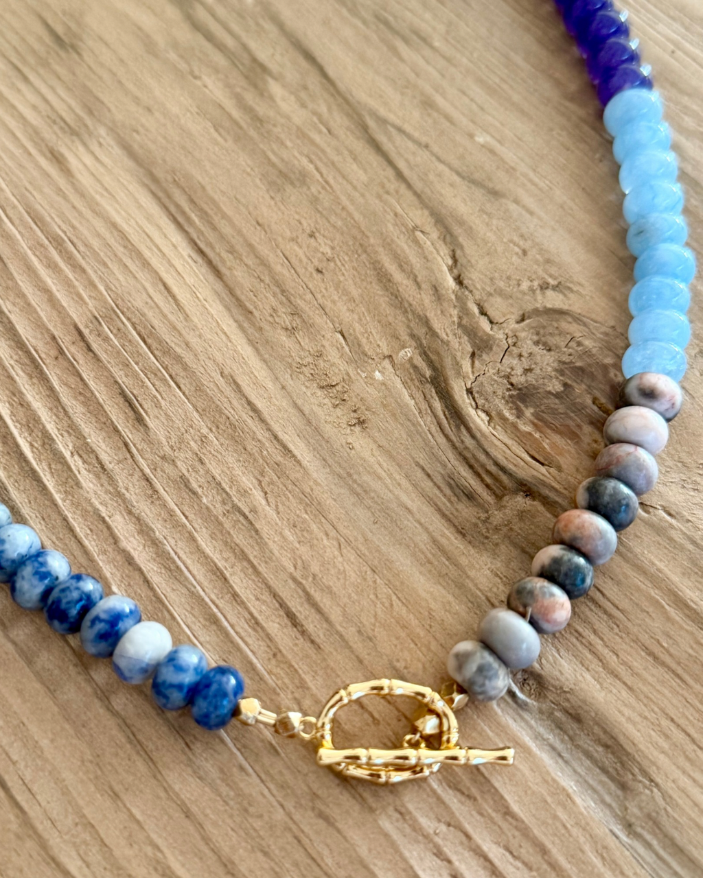Close-up of Violet Hour handmade necklace showing blue marbled jasper, soft aqua and neutral gemstone beads with textured gold toggle clasp.
