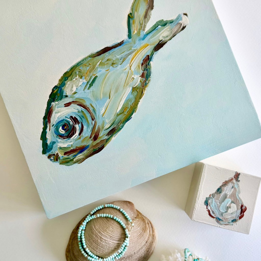 An abstract painted fish artwork on a square canvas, styled on a white surface with a small matching painted box, a seashell with turquoise bead jewelry, and soft decorative props.