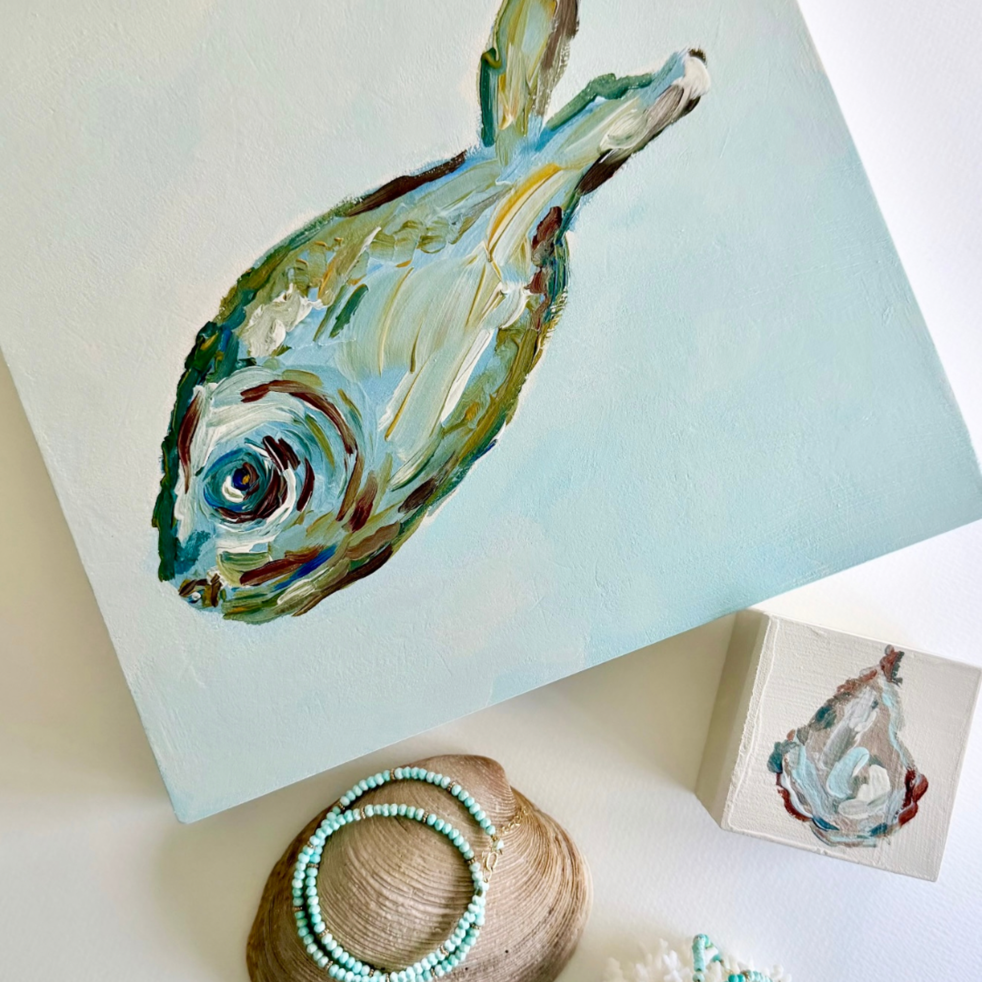 An abstract painted fish artwork on a square canvas, styled on a white surface with a small matching painted box, a seashell with turquoise bead jewelry, and soft decorative props.