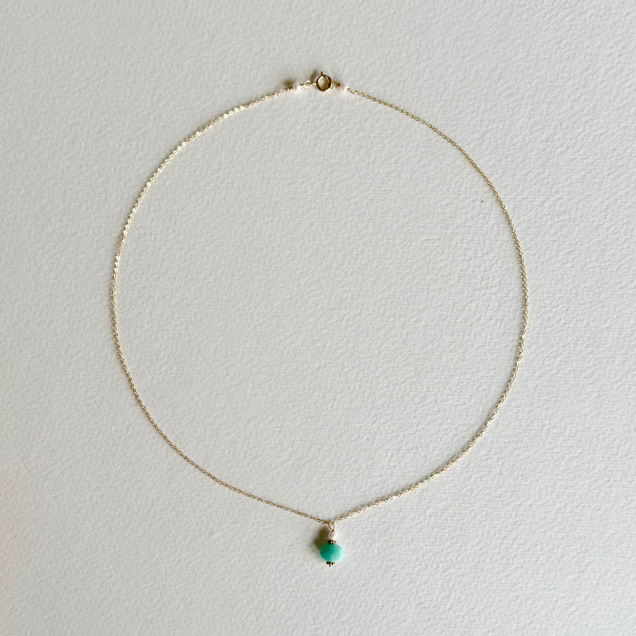 A delicate gold chain necklace with a small, round turquoise bead pendant, displayed on a textured white background.