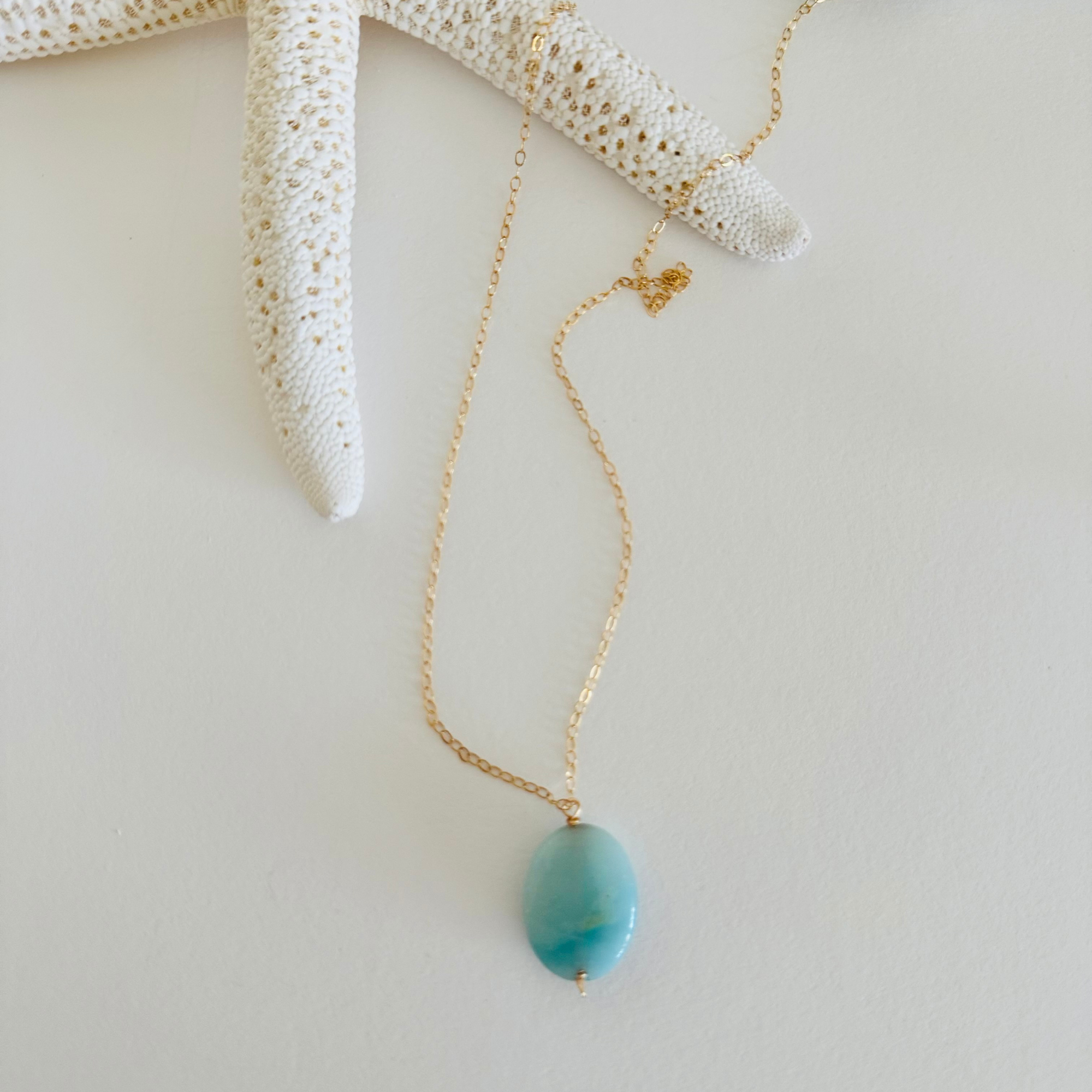 Aquamarine sea drop pendant with light blue color variation on 14k gold filled wire chain draped across a creamy white starfish.