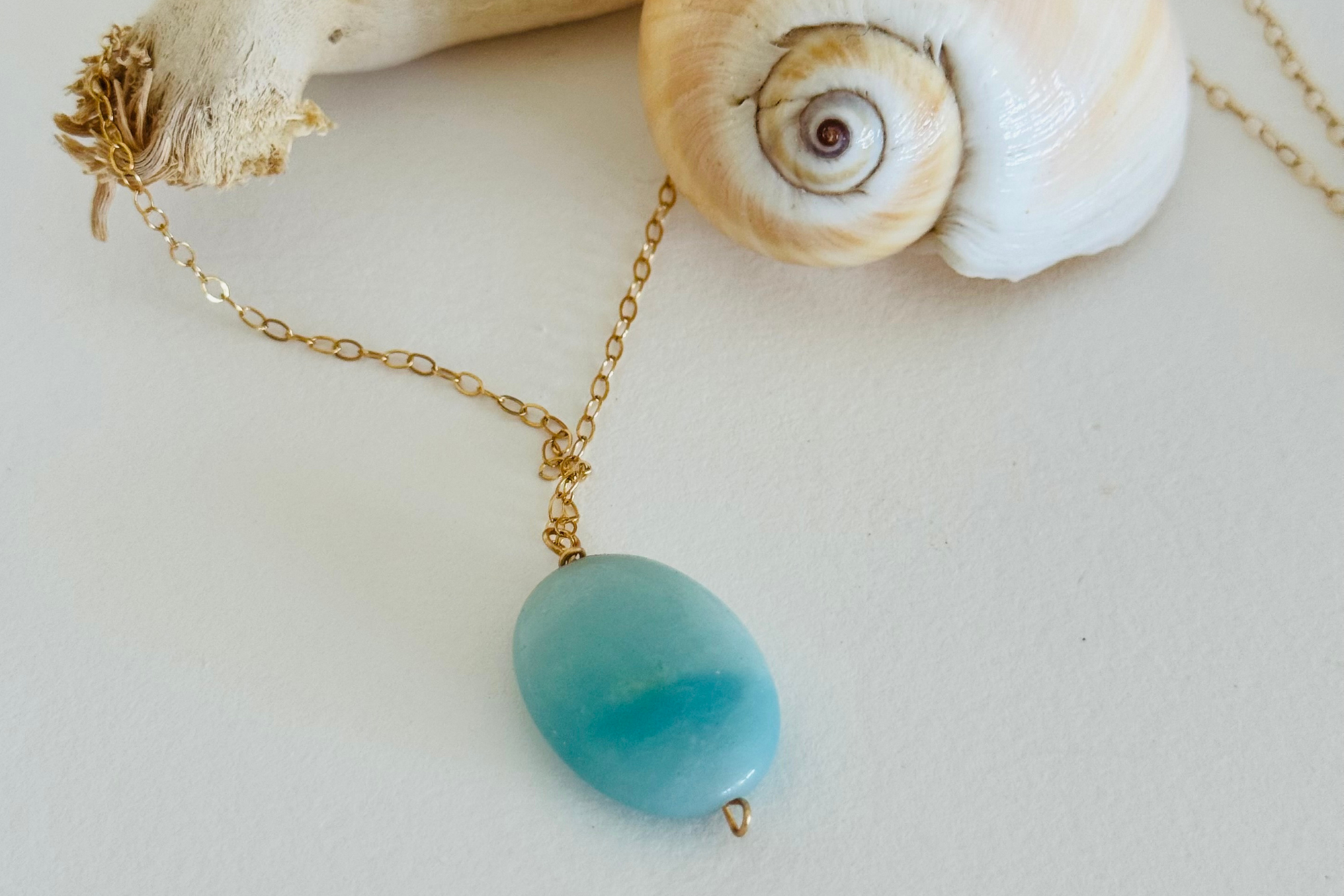 Aquamarine sea drop pendant with light blue color variation on 14k gold filled wire chain draped across a spiral seashell and bleached driftwood branch.