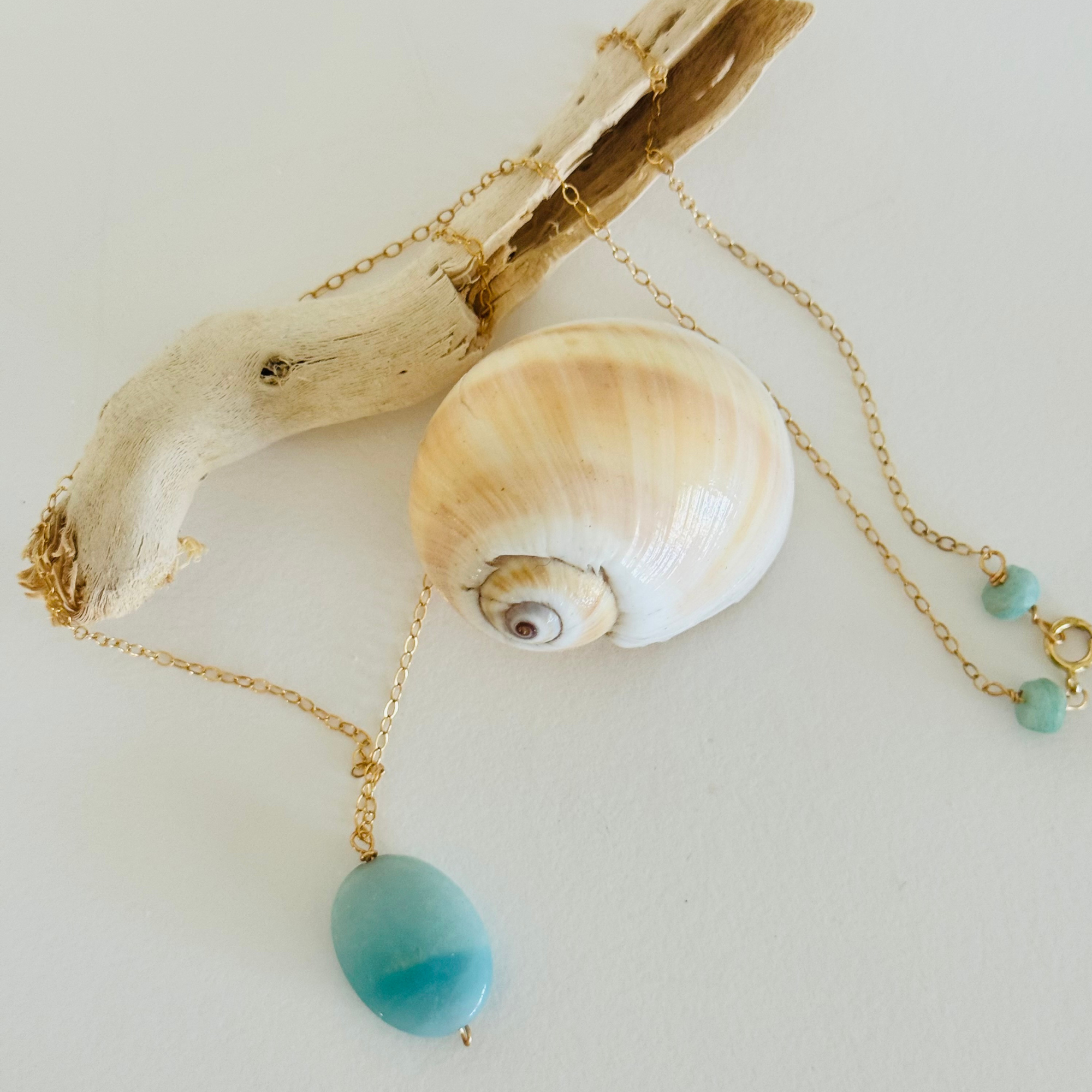 Aquamarine sea drop pendant with light blue color variation on 14k gold filled wire chain draped across a spiral seashell and bleached driftwood branch from aerial view. Two seafoam beads offset the clasp.
