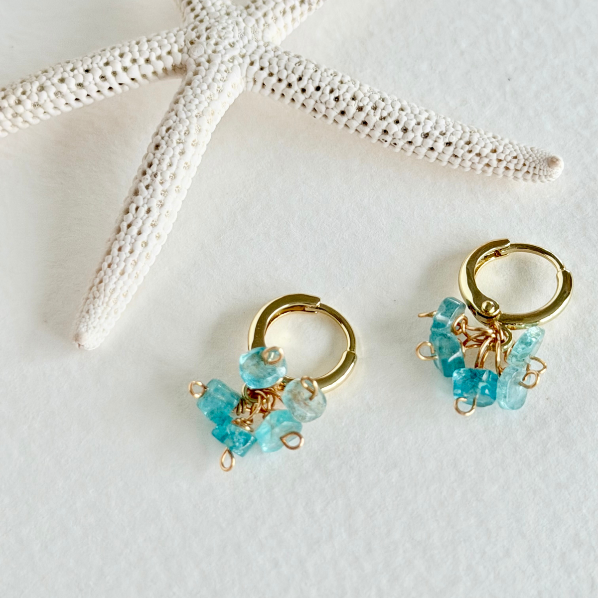 Gold hoop earrings with aquamarine beads on a white surface with a starfish.