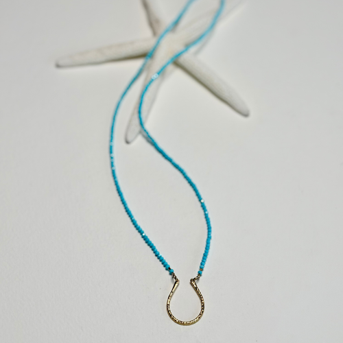 A delicate turquoise beaded necklace with a small gold horseshoe pendant, styled on a white surface with a white starfish prop.