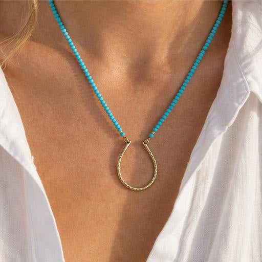 A close-up of a person wearing a necklace with small turquoise-blue beads and a textured gold hoop pendant, styled with a white collared shirt.
