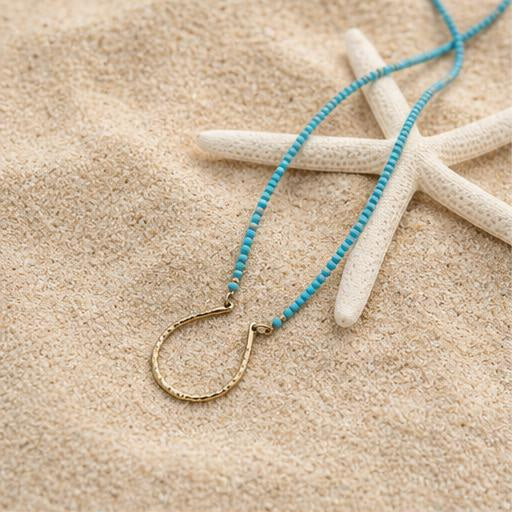 A turquoise beaded necklace with a textured gold-tone circular pendant, styled on beige sand next to a white starfish.