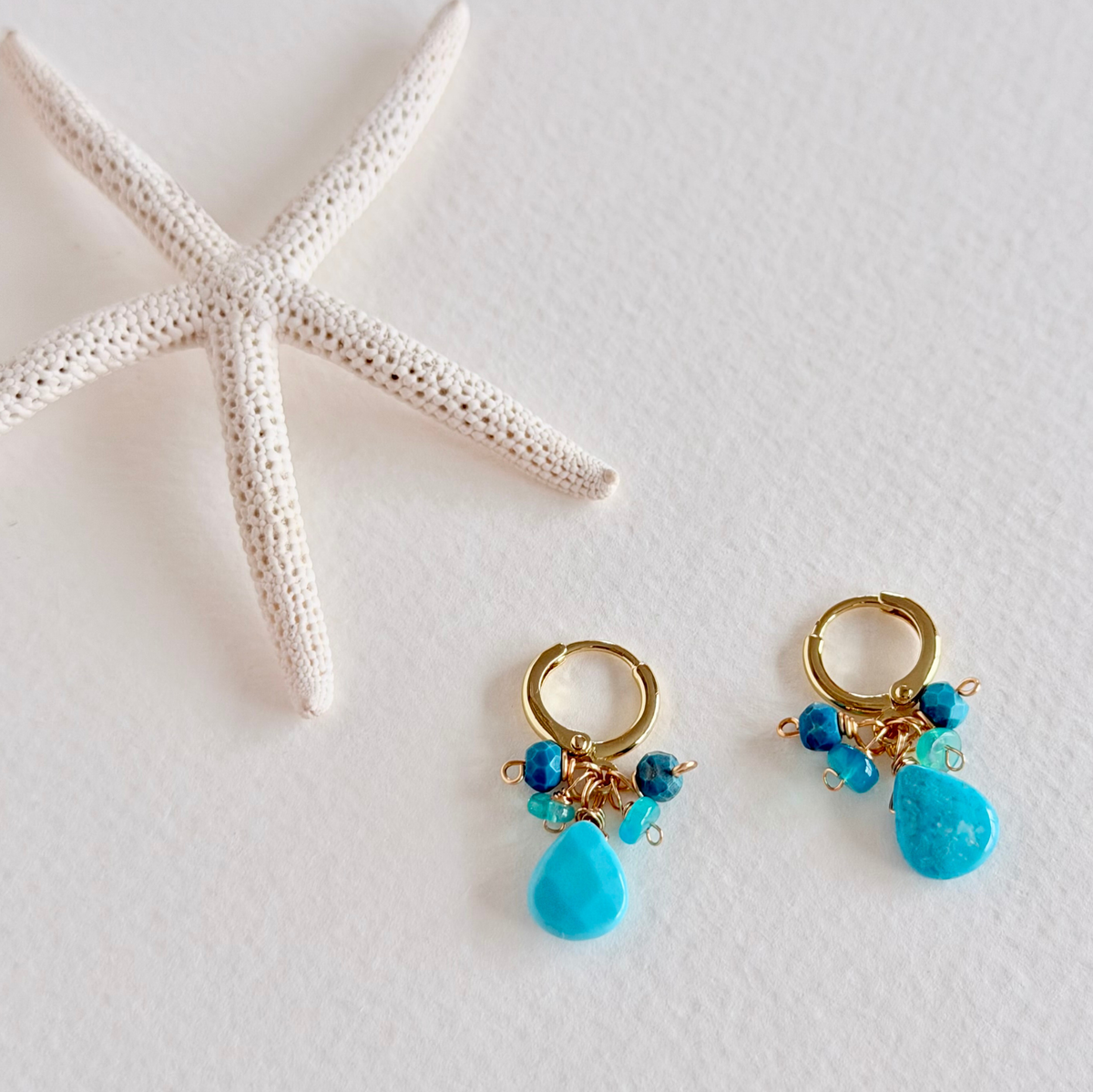 Gold hoop earrings with blue teardrop beads on a white background with a starfish.