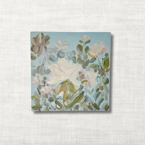 A square canvas art print featuring a painted floral design with white, pink, and green flowers on a light blue background, displayed against a textured off-white linen backdrop.