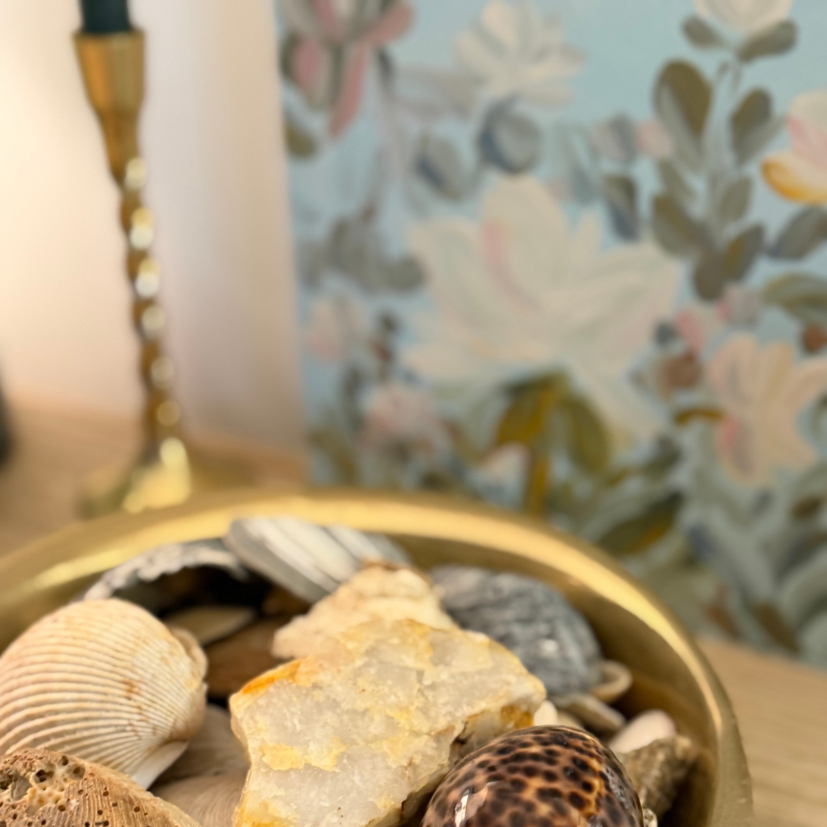 A close-up of a gold-toned bowl filled with assorted seashells and a rough crystalline stone, set against a softly blurred floral wallpaper background.