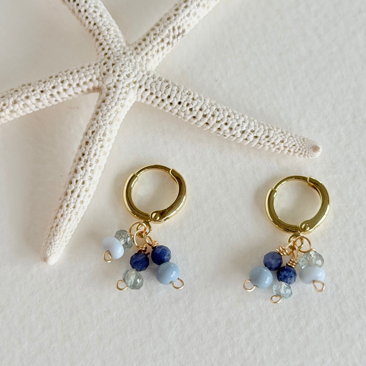 Gold hoop earrings with blue and green bead charms on a white surface with starfish.