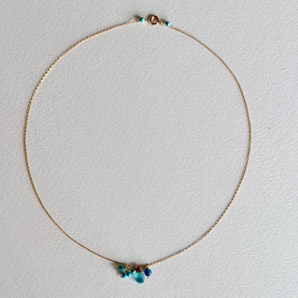 A delicate gold chain necklace with small turquoise blue beads and a small cluster of blue and purple stones at the center, displayed on a textured white background.
