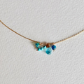 A delicate gold chain necklace featuring a small cluster of beads in shades of blue, turquoise, and teal, displayed on a textured white background.