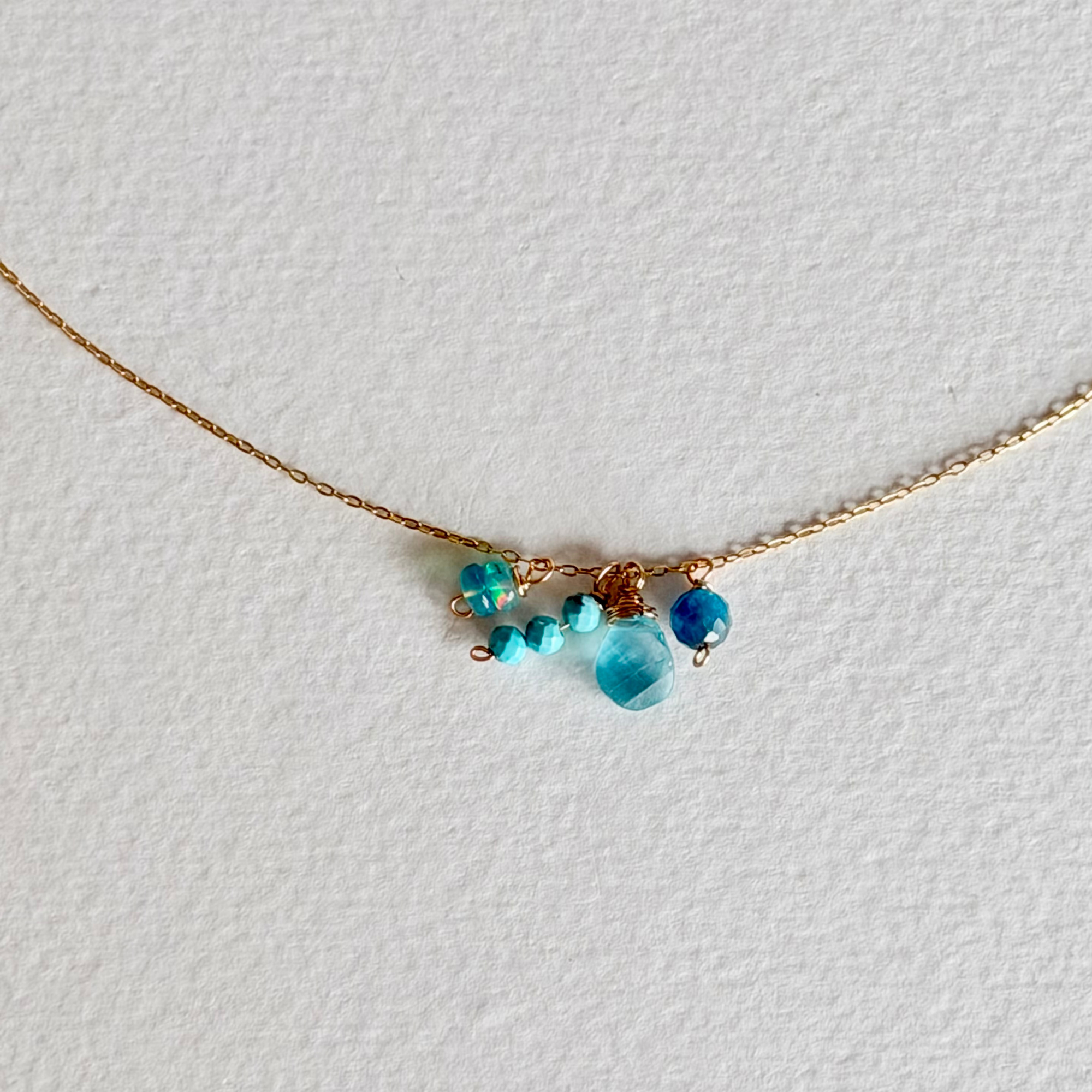 A delicate gold chain necklace featuring a small cluster of beads in shades of blue, turquoise, and teal, displayed on a textured white background.