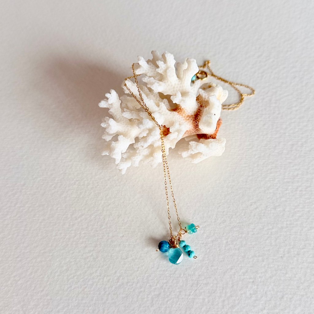 A delicate gold chain necklace with turquoise blue beads, displayed draped over a white coral shell on a textured off-white background.