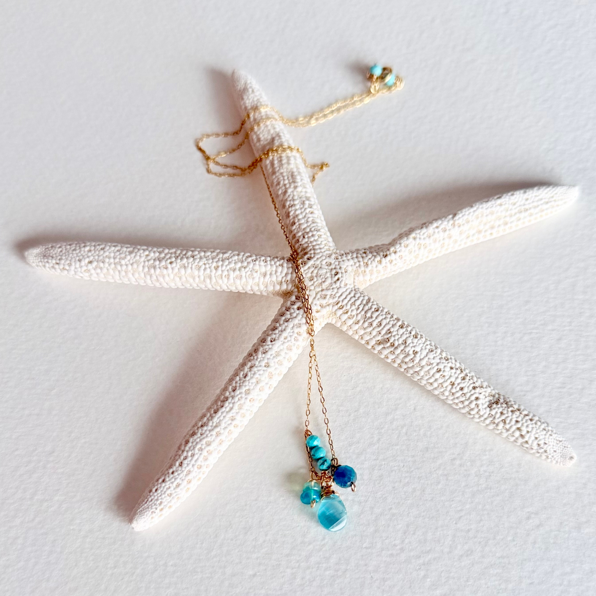A delicate gold chain necklace with turquoise and blue beads draped over a white starfish prop on a textured white background.