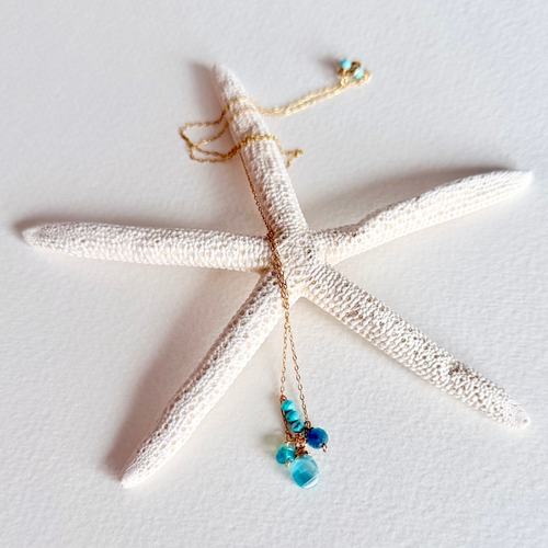 A delicate gold chain necklace with turquoise and blue beads draped over a white starfish prop on a textured white background.