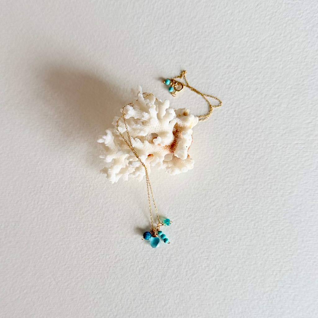 A delicate gold necklace with small turquoise blue beads and a white coral shell pendant, displayed on a textured white background.