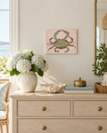 A square canvas art print featuring a blue-green crab on a soft pink background, styled in a bright, coastal-themed room above a light wood dresser with a vase of white hydrangeas and other home decor.