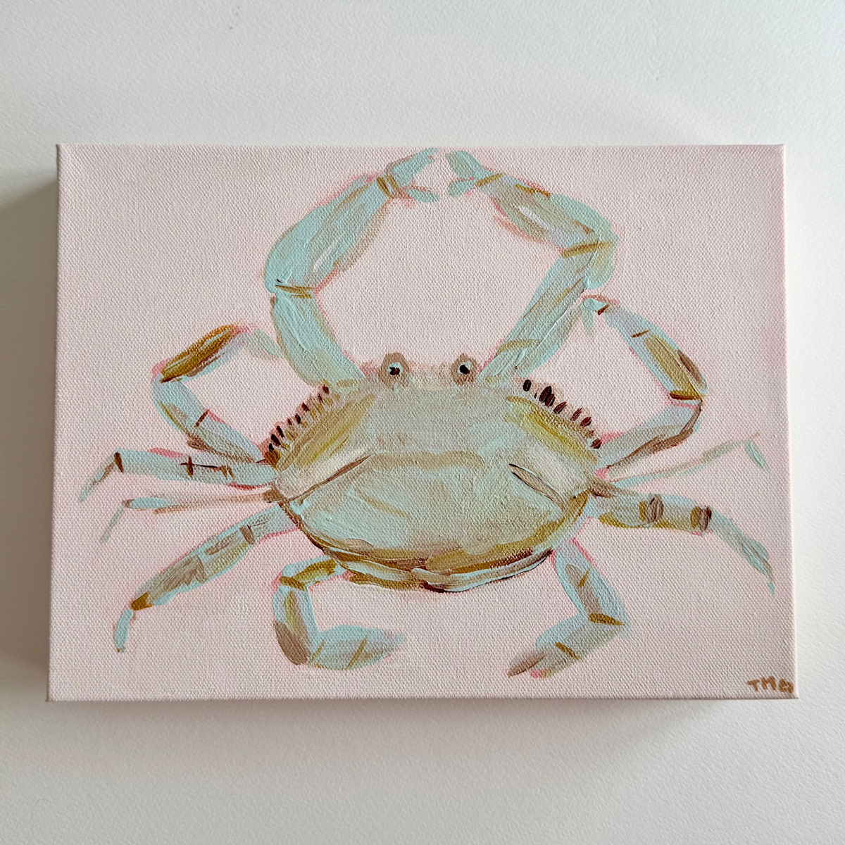 A rectangular acrylic canvas painting of a light blue-green crab on a pale pink background, mounted on a white wall.