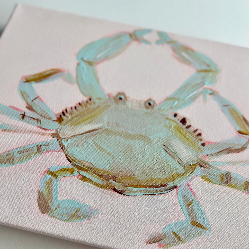 A close-up of a canvas painting featuring a light blue and beige crab on a pale pink background.