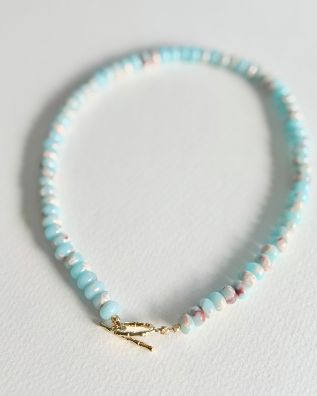 A beaded bracelet with light blue, white, and pink marbled beads, finished with a gold toggle clasp, displayed on a light gray textured surface.