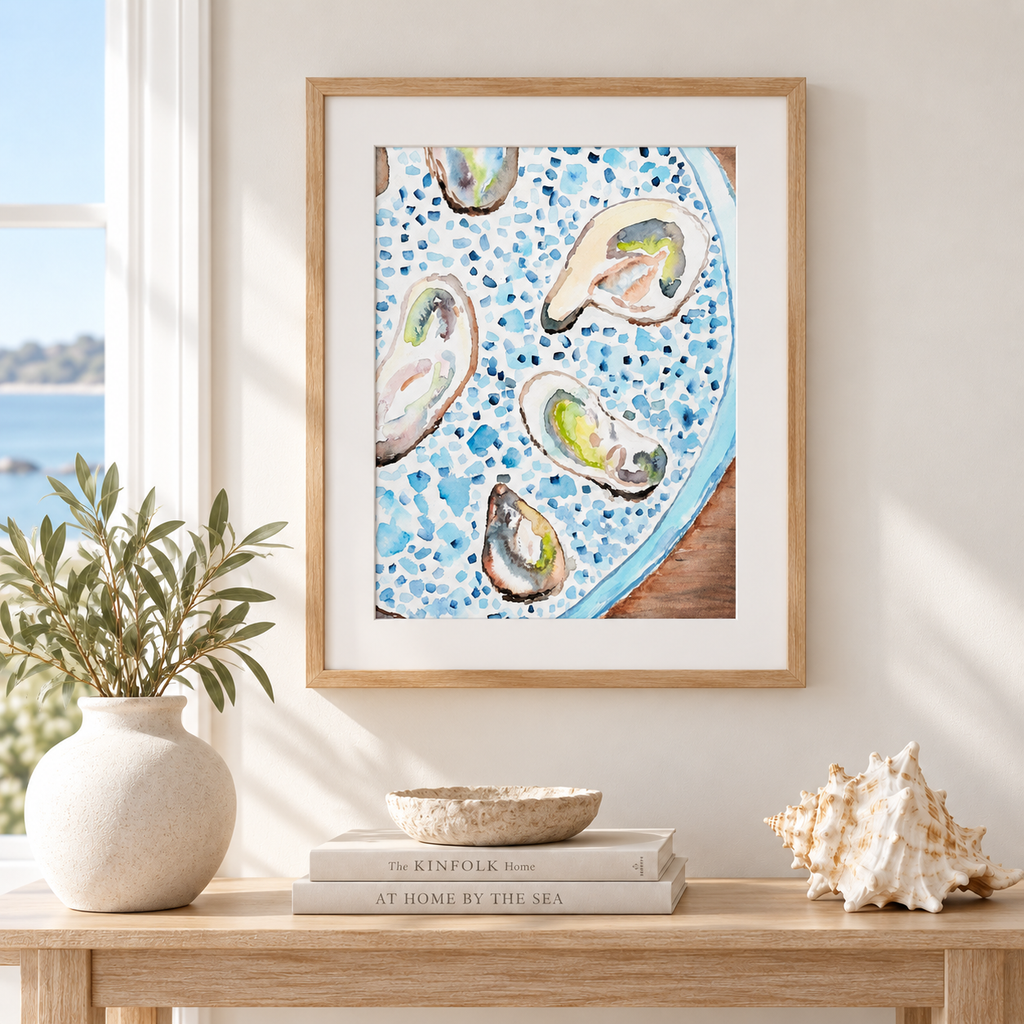 Framed wall art featuring a watercolor-style plate of oysters on a speckled blue plate, displayed in a light wood frame on a beige wall above a wooden console table with a vase, books, and a seashell.
