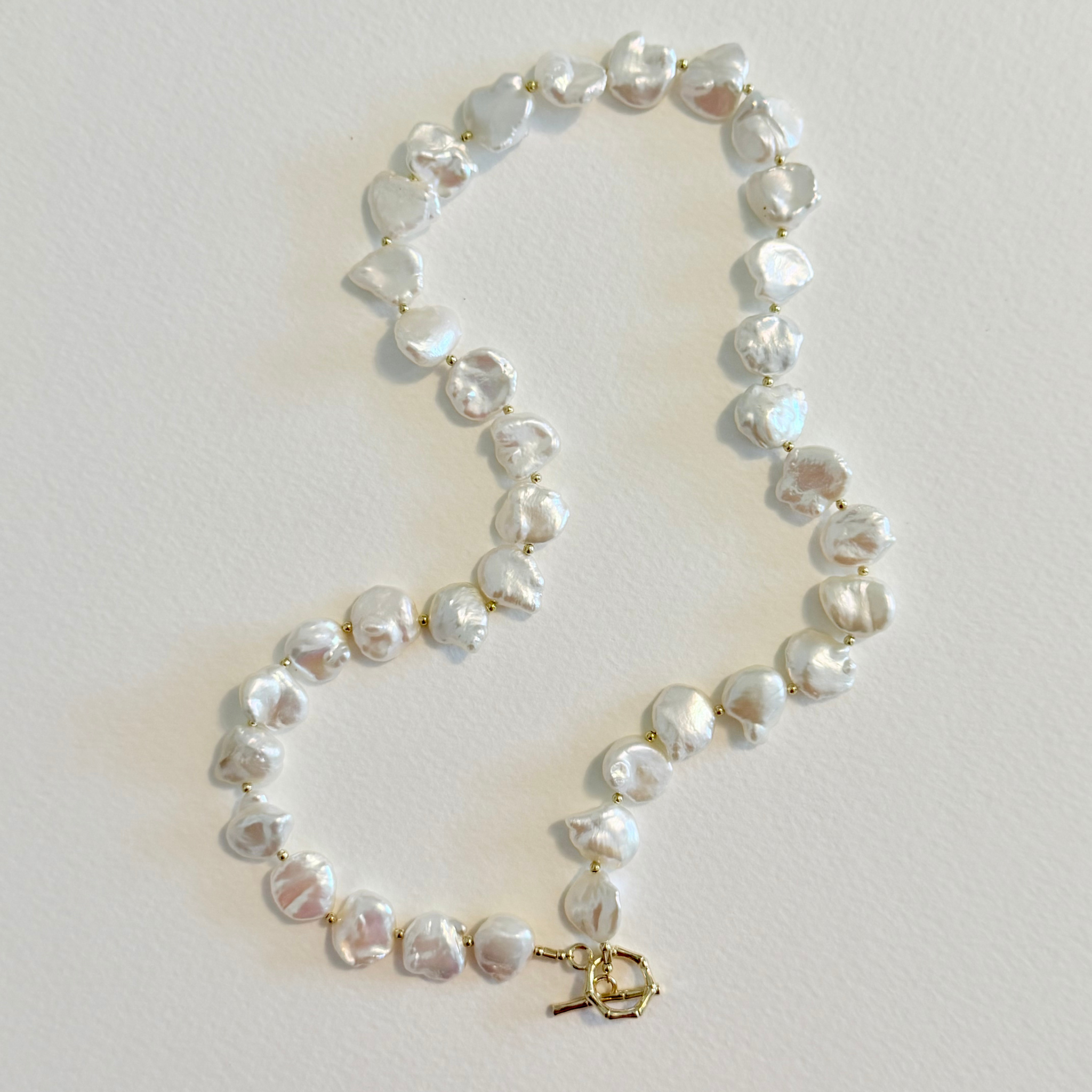 A strand of irregular white baroque pearls linked with small gold-toned metal findings, including a gold toggle clasp, laid out on a light beige textured background.