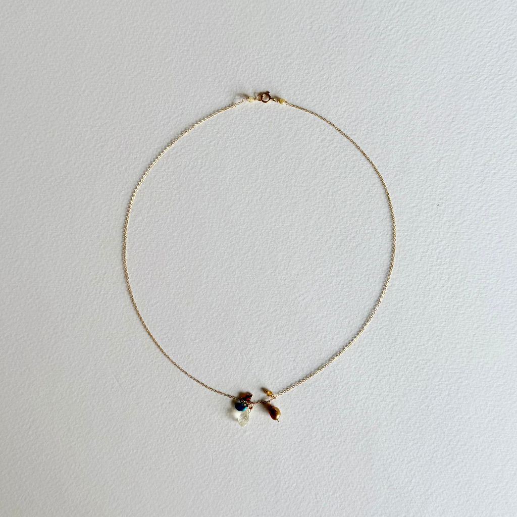 A delicate gold chain necklace with a small bow charm and a tiny blue bead accent, displayed on a textured white background.