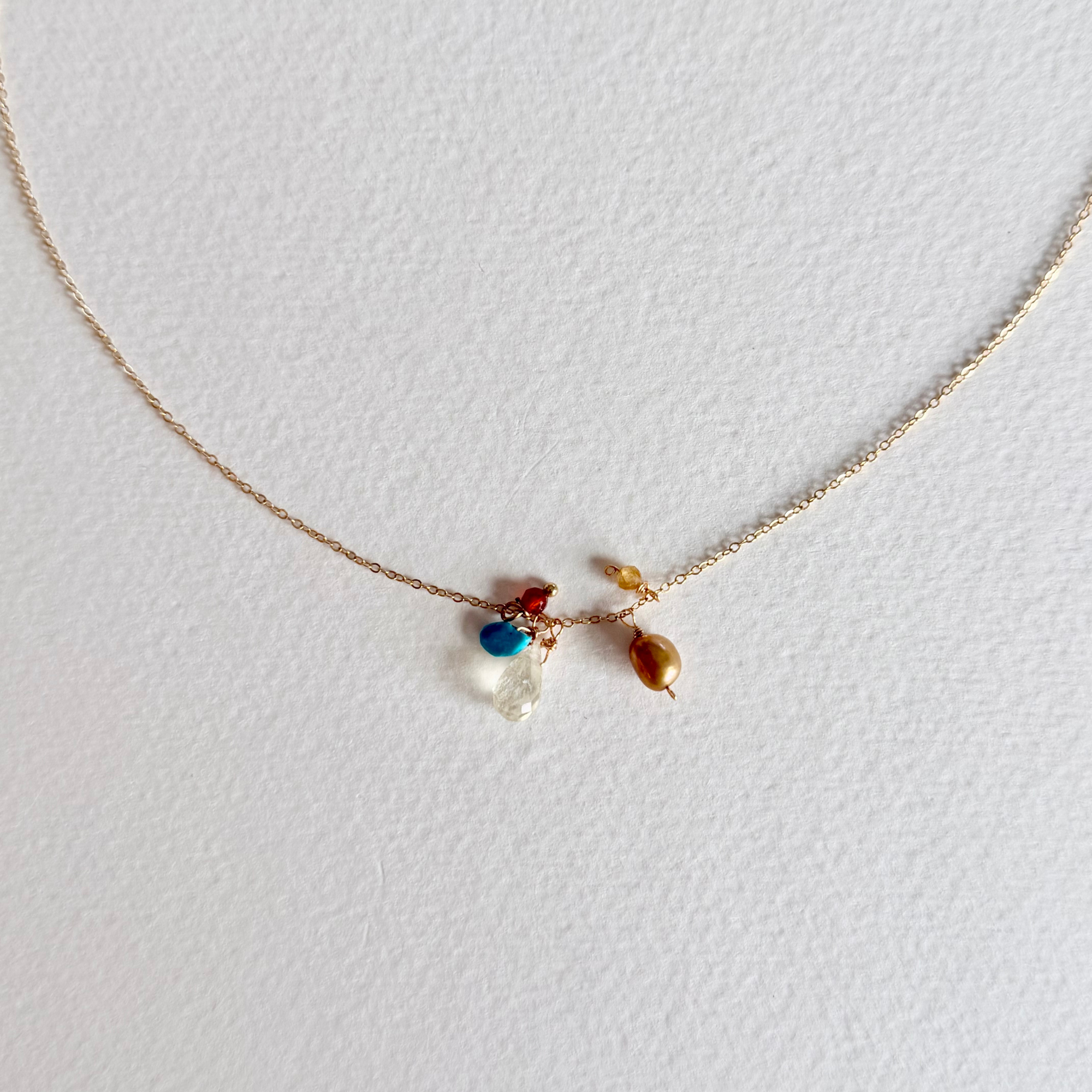 A delicate rose gold chain necklace with a small central cluster of charms, including a blue bead, a clear faceted stone, and a brown teardrop bead, displayed on a textured white background.