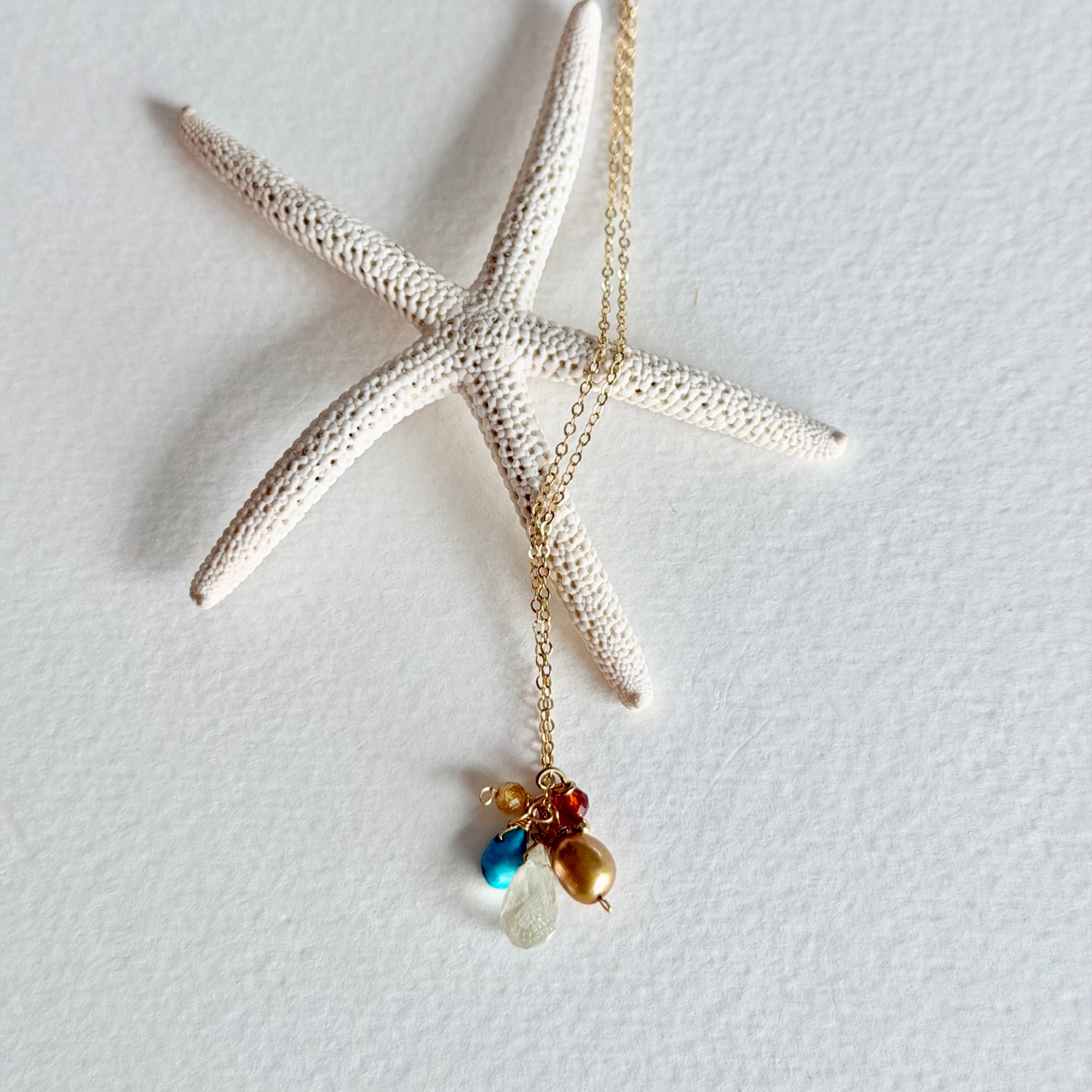A gold chain necklace with a starfish-shaped pendant and small colorful bead charms, displayed on a textured white background with a white starfish prop.