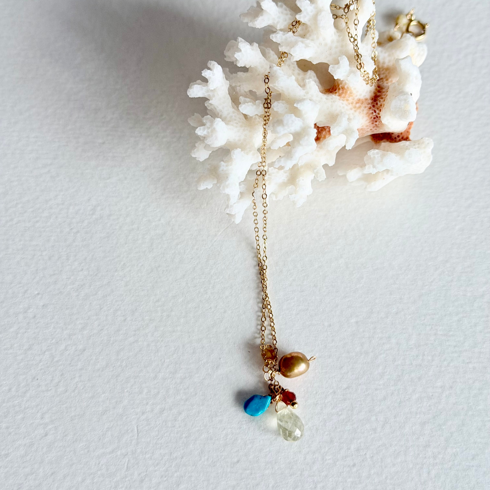 A delicate gold chain necklace with a drop pendant featuring a turquoise blue bead, a small red bead, a gold bead, and a clear faceted stone, displayed on a white coral shell against a white background.