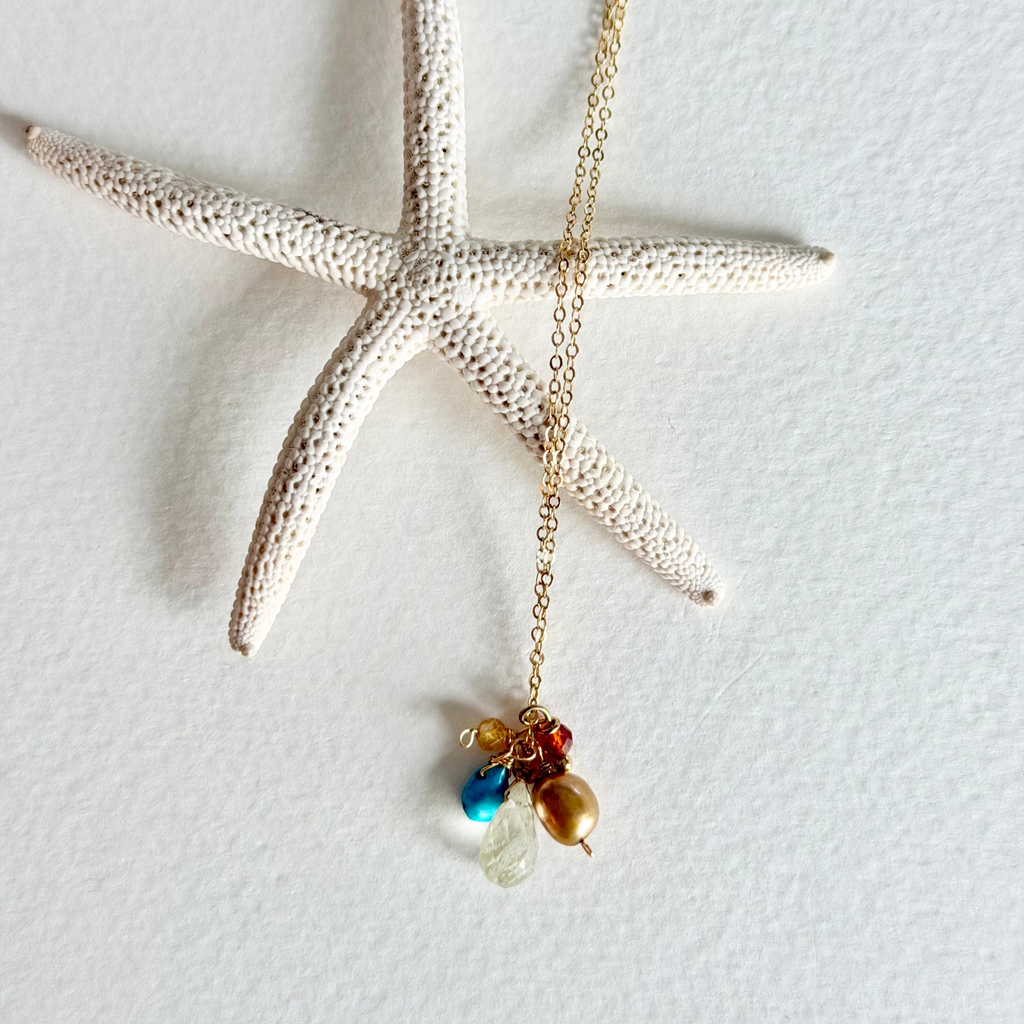 A gold chain necklace with a small cluster of colorful beads, including blue, red, brown, and clear teardrop stones, displayed on a textured white background with a decorative white starfish prop.
