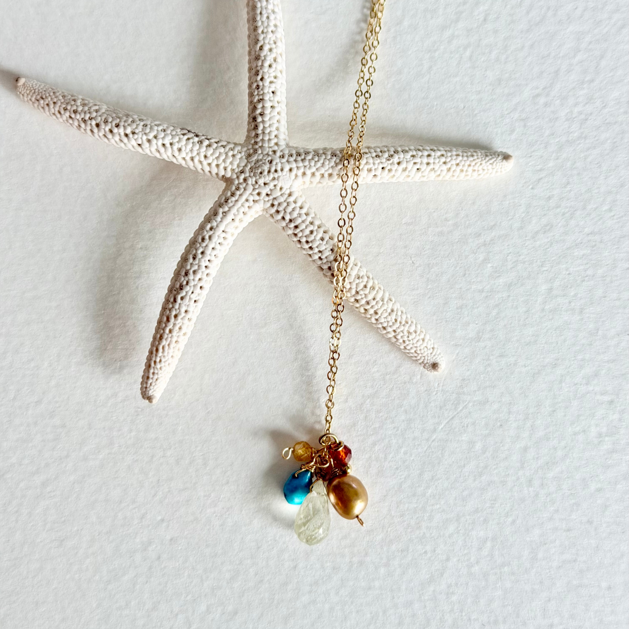 A gold chain necklace with a small cluster of colorful beads, including blue, red, brown, and clear teardrop stones, displayed on a textured white background with a decorative white starfish prop.