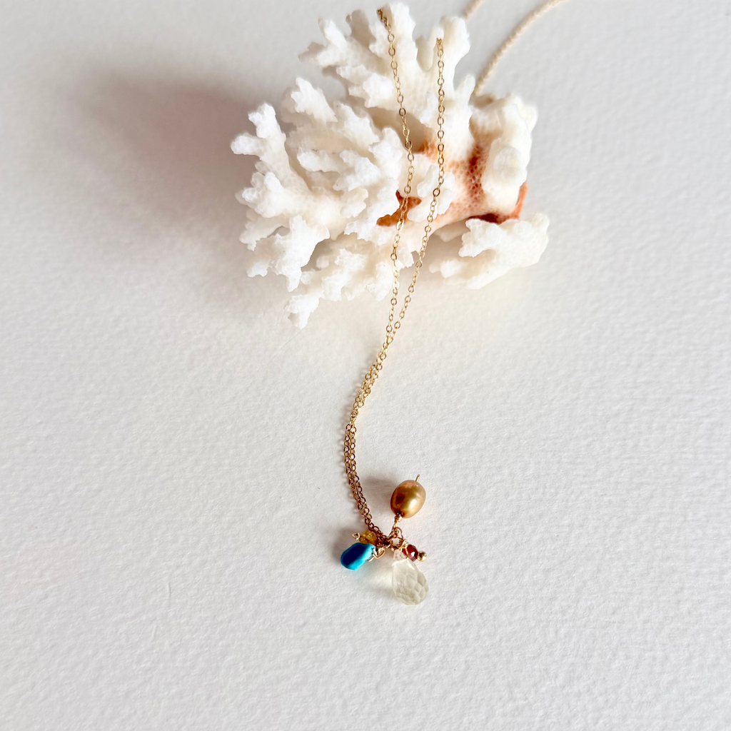 A delicate gold necklace with a pendant featuring a gold bead, a small blue bead, and a clear glass heart, displayed on a white coral shell against a white background.