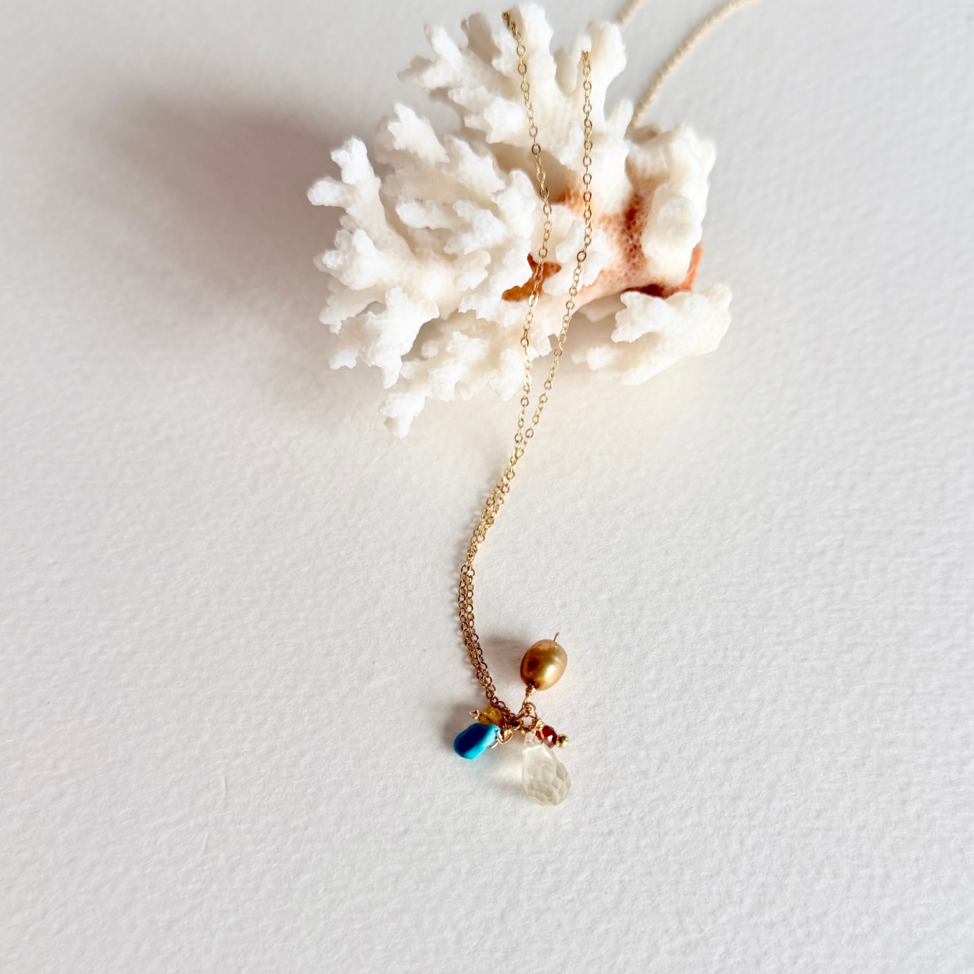 A delicate gold necklace with a pendant featuring a gold bead, a small blue bead, and a clear glass heart, displayed on a white coral shell against a white background.