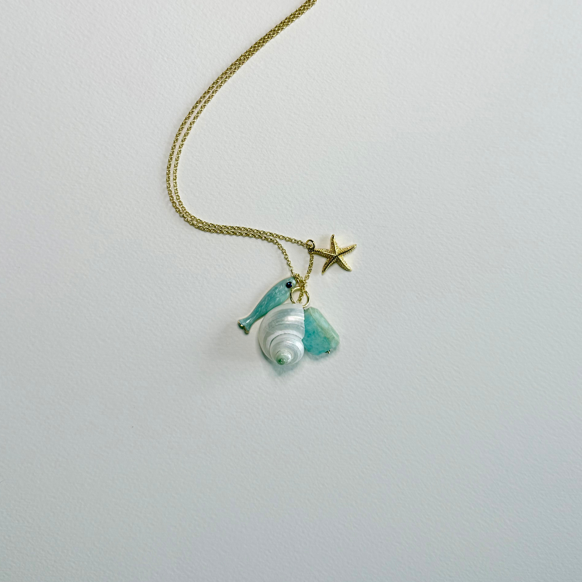 A delicate gold chain necklace featuring a starfish charm, a small blue dolphin charm, and a white seashell charm, displayed on a clean white background.