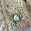 A close-up of a handmade seashell and starfish brooch pinned onto a chunky cream cable-knit sweater. The brooch features a white seashell, a turquoise ribbon, a turquoise fish, a gold starfish, and pearl-like beads.