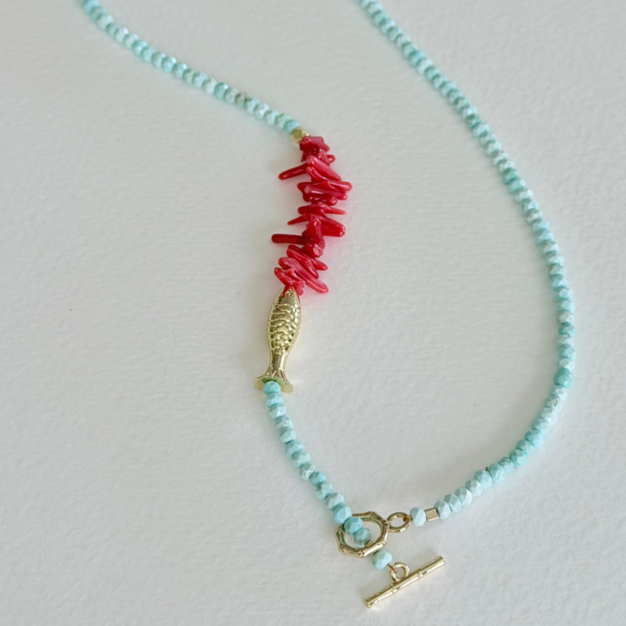 Close up of Handmade faceted aqua turquoise necklace with natural coral centerpiece, gold fish charm, and 14k gold-filled bamboo toggle clasp by Marie & Oakley.