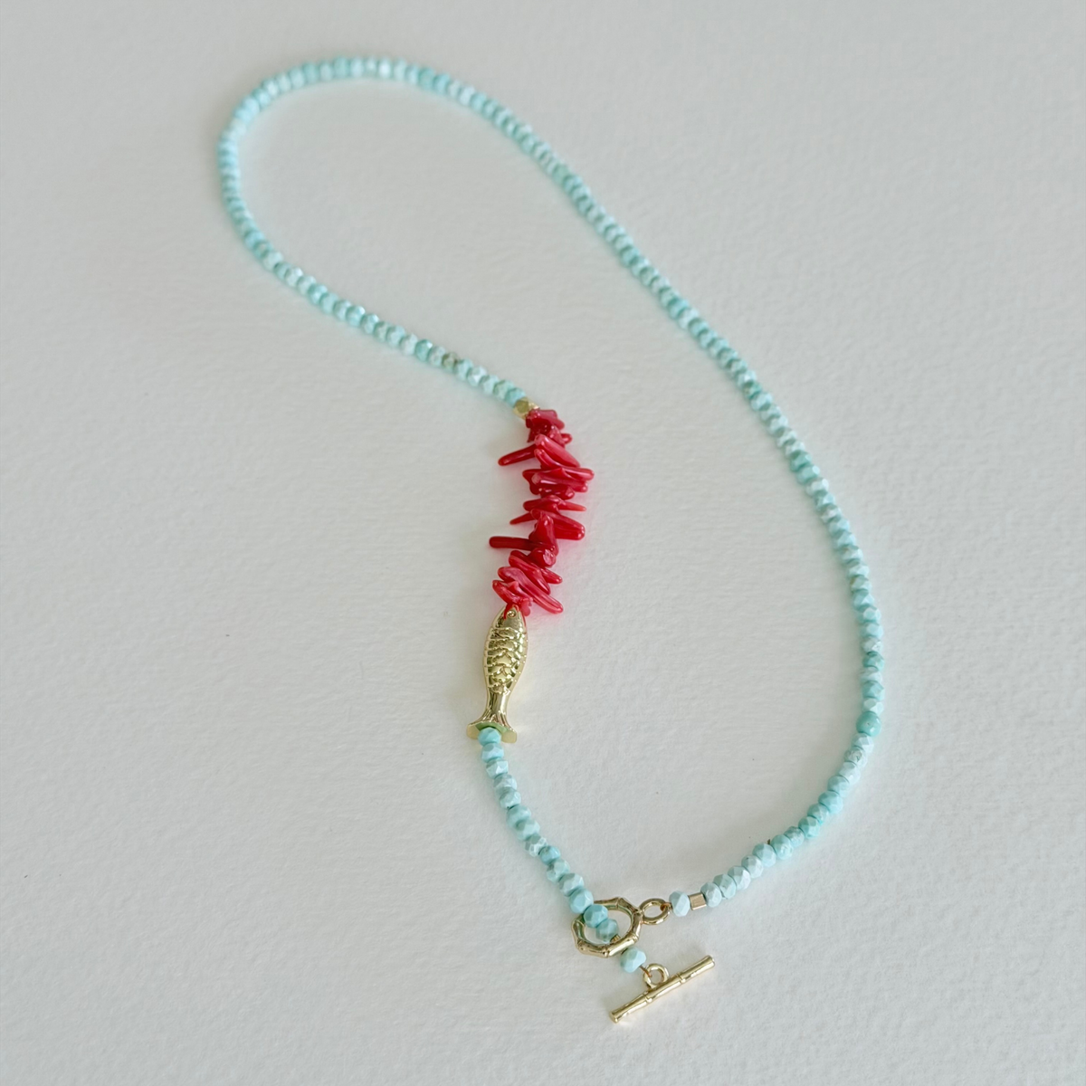 Handmade faceted aqua turquoise necklace with natural coral centerpiece, gold fish charm, and 14k gold-filled bamboo toggle clasp by Marie & Oakley.