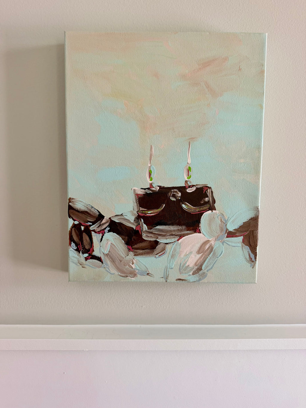 An abstract canvas painting hanging on a beige wall, featuring a dark brown crab with two thin vertical elements and soft beige, white, and pale blue brushstrokes around it.