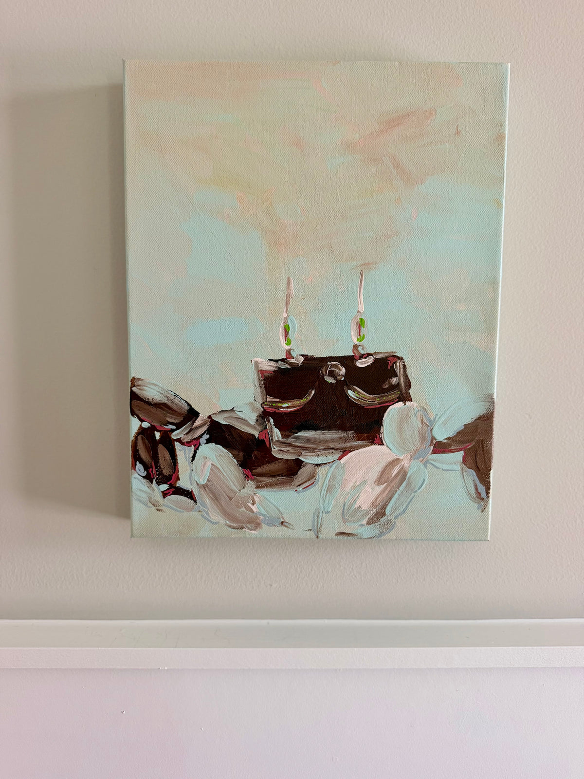 An abstract canvas painting hanging on a beige wall, featuring a dark brown crab with two thin vertical elements and soft beige, white, and pale blue brushstrokes around it.