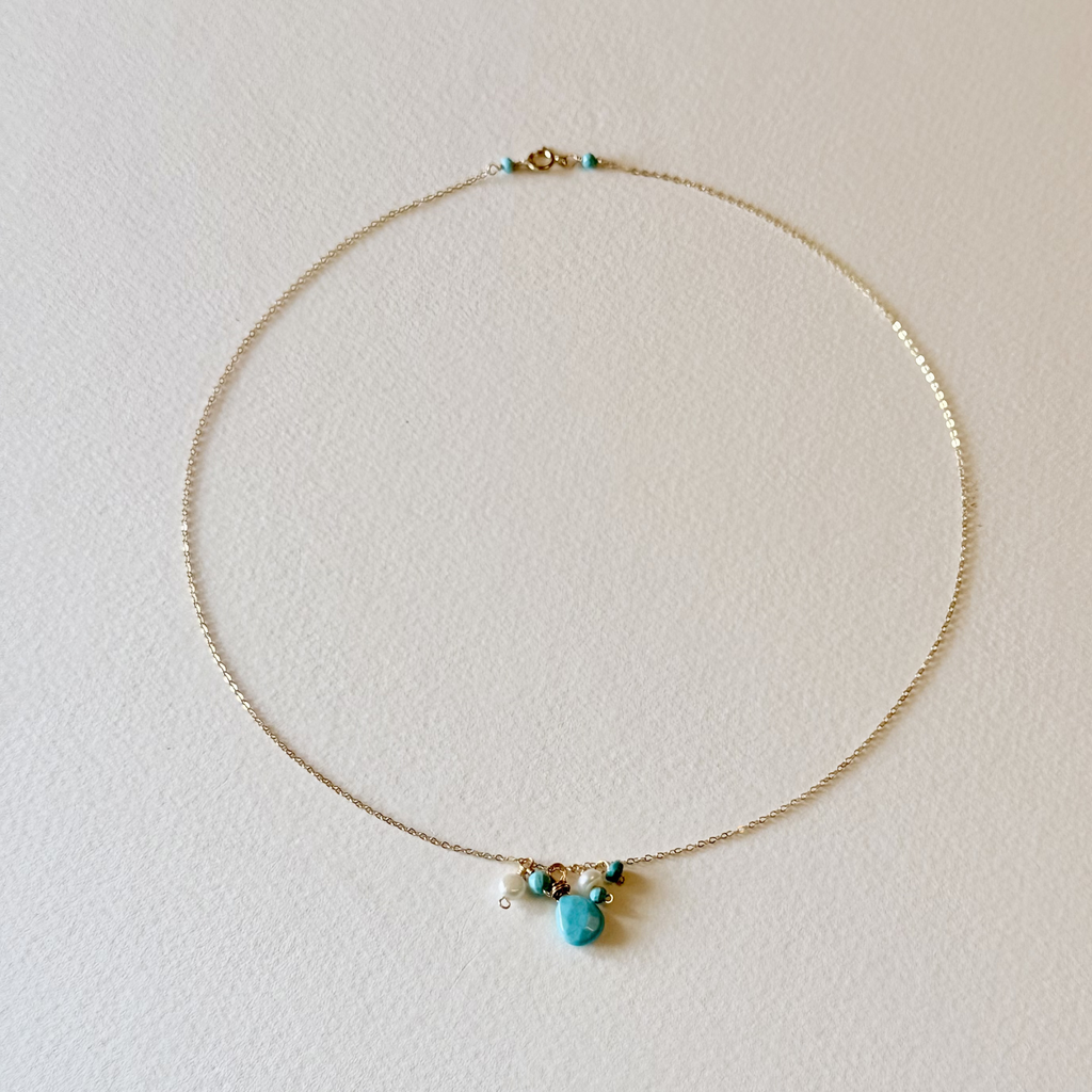 A delicate gold chain necklace with a small cluster of turquoise and gold beads at the center, displayed on a textured off-white background.