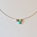 A delicate gold chain necklace featuring a small cluster of turquoise and white beads with a central turquoise heart charm, displayed on a textured off-white background.