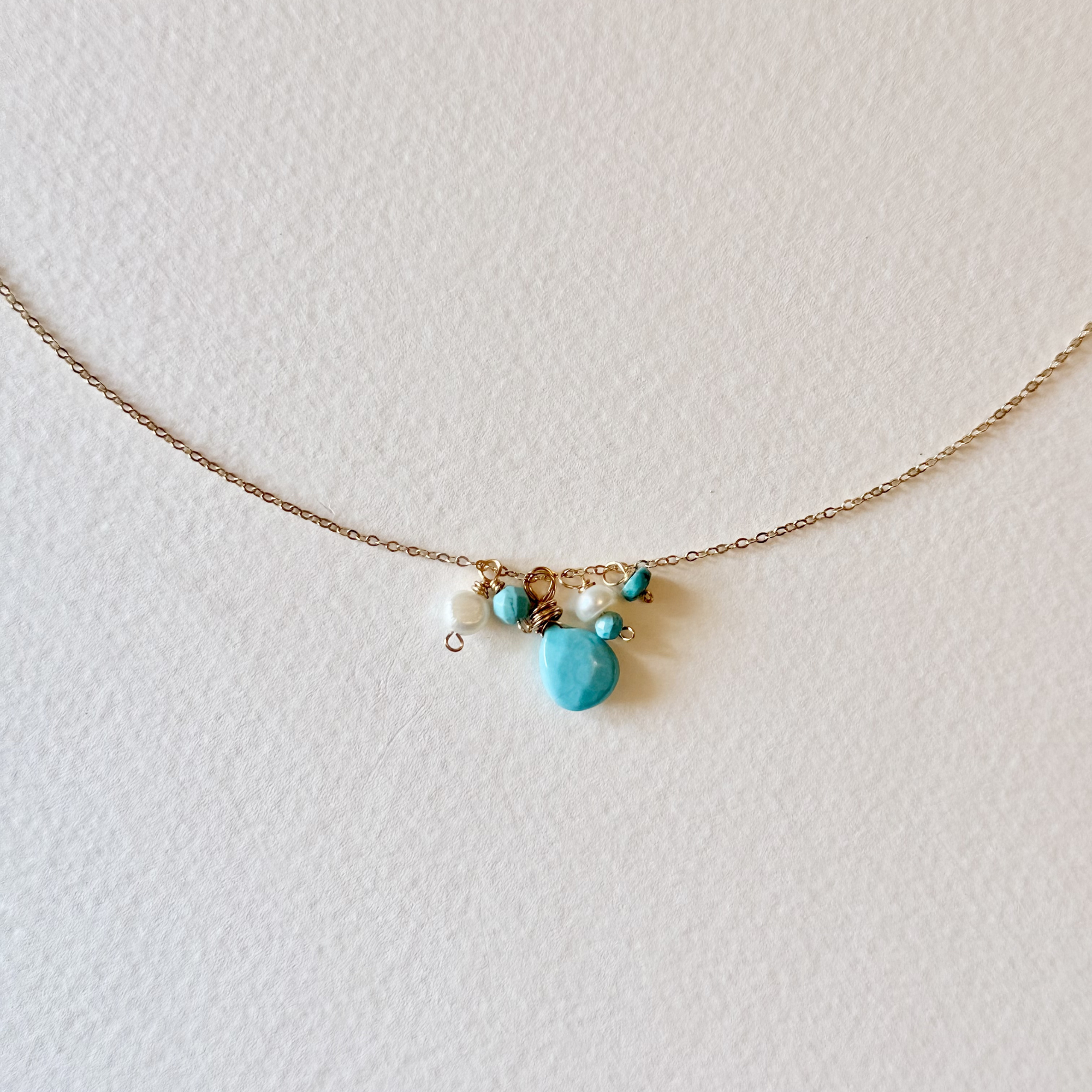 A delicate gold chain necklace featuring a small cluster of turquoise and white beads with a central turquoise heart charm, displayed on a textured off-white background.