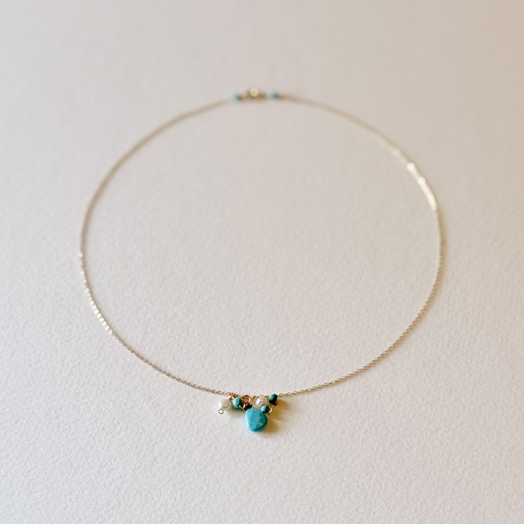 A delicate gold chain necklace with a small cluster of turquoise blue beads and a central turquoise stone pendant, displayed on a textured off-white surface.