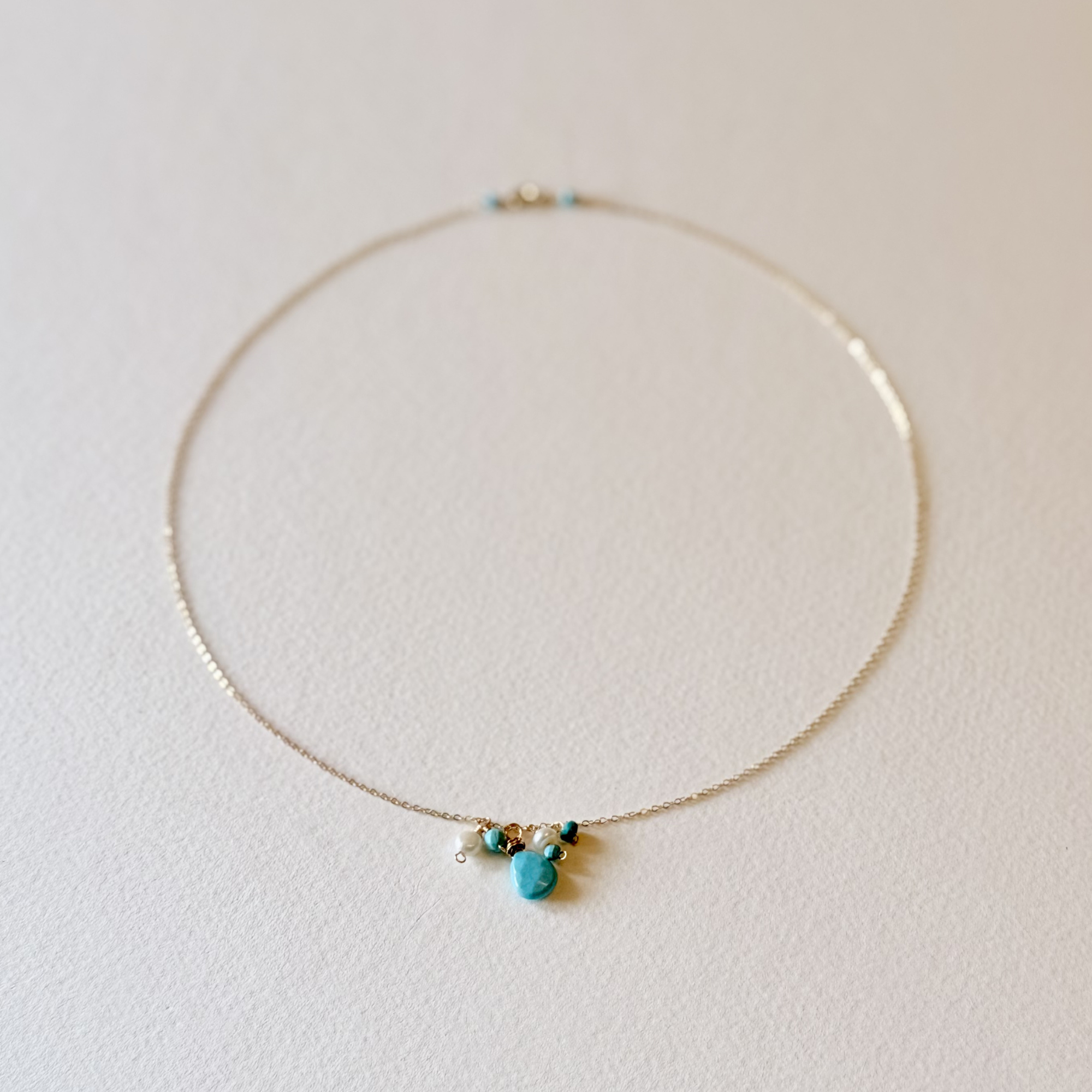A delicate gold chain necklace with a small cluster of turquoise blue beads and a central turquoise stone pendant, displayed on a textured off-white surface.